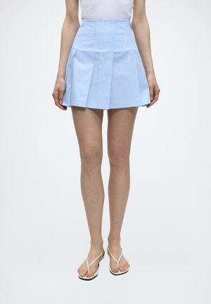 Kobieta nosząca jasnoniebieską plisowaną mini spódnicę z cienkimi białymi paskami, biały bezrękawnik i beżowe sandały na obcasie.