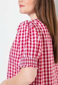 Blouse à carreaux rouges et blancs avec des manches bouffantes, des poignets smockés, et un tissu en coton léger, offrant une coupe décontractée.