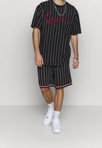 Czarna, szeroka koszulka w paski z czerwonym napisem, zestawiona z pasującymi czarnymi szortami z czerwonymi i białymi akcentami oraz białymi trampkami.