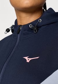 Mornarska zip-up dukserica s ružičastim logotipom, podesivim vezicama oko vrata, glatkom tkaninom, kontrastnim sivo panelom i metalnim elementima.