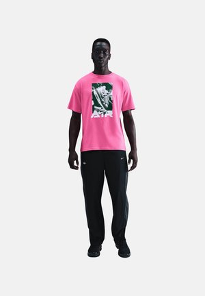 Homme debout portant un t-shirt rose vif avec un graphique de sneaker et le mot Air, pantalon noir et baskets noires sur fond blanc.