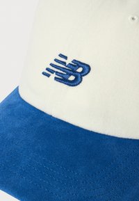 Balta ir mėlyna kepurė su mėlynai siuvinėtu stilizuotu "NB" logotipu priekinėje panelėje ir mėlyna suede kreiva briauna.