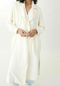 Dlouhý béžový trench coat s klopovým límcem, páskem v pase, bočními rozparky a hladkou strukturou. Model má v ruce stříbrnou sponu.