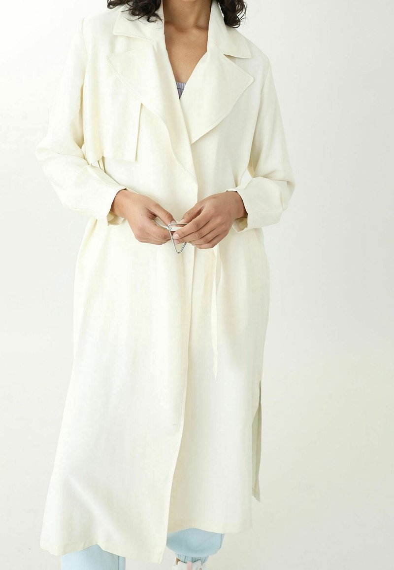 Dlouhý béžový trench coat s klopovým límcem, páskem v pase, bočními rozparky a hladkou strukturou. Model má v ruce stříbrnou sponu.
