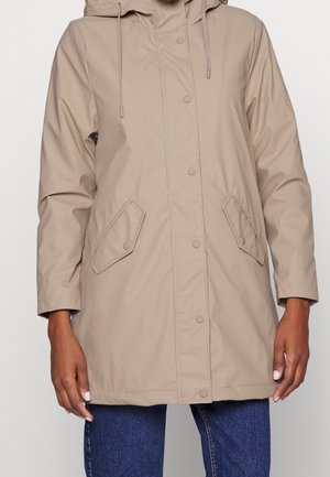 Beige huva regnjacka med tryckknappar, sidofickor och en något lös passform. Slät textur och vattentåligt material.