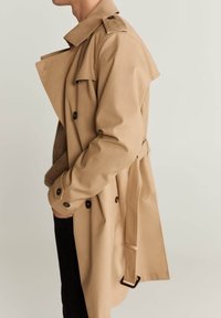 Beige Trenchcoat mit doppelreihigem Design, Schulterklappen und einem Gürtel. Das Material wirkt glatt mit einem leichten Glanz.