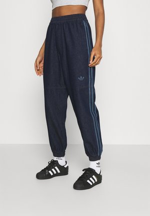 Personne portant un pantalon de sport Adidas bleu foncé avec des bandes bleu clair, des chaussettes blanches Adidas et des baskets noires Adidas, debout sur un sol clair.