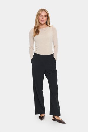 Beige Langarmoberteil mit glatter Textur, kombiniert mit schwarzen, weit geschnittenen Hosen mit Fronttaschen und einem gefalteten Design. Dunkle Schuhe.