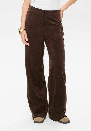 Braune Corduroy-Hosen mit weitem Bein und strukturierter Oberfläche, mit hohem Bund und subtiler vertikaler Rippung, kombiniert mit beige-farbenem Schuhwerk.