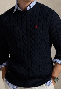 Navy cable-knit sweater with ribbed hem and cuffs, featuring a small red logo. Worn over a blue and white striped collared shirt.