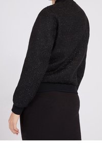 Chaqueta negra con textura, con una cintura y puños de canalé ajustados. Hilos brillantes tejidos a lo largo para un brillo sutil.