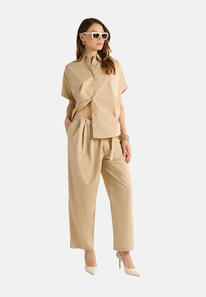 Camicia button-up a maniche corte color beige in cotone con design cropped, abbinata a pantaloni beige larghi. Scarpe col tacco alte bianche e occhiali da sole.