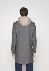 Cappotto grigio in misto lana con cappuccio imbottito beige. Presenta una vestibilità sartoriale, maniche lunghe e un design minimalista. Vista dal retro visibile.