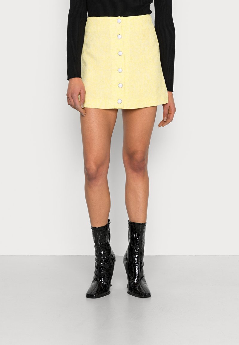 Mini-jupe jaune en tissu texturé, avec un bouton sur le devant et une finition lisse. Associée à des bottines noires brillantes.