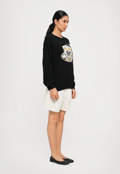 Schwarzer Pulli mit langen Ärmeln und gesticktem Teetassenmotiv, kombiniert mit weißen Shorts und schwarzen flachen Schuhen, vor einem schlichten Hintergrund.