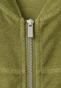 Close-up of silver zipper on green textured fabric, showing zipper teeth and pull tab in a centered, vertical position.