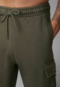 Olivengrüne Sweatpants mit Kordelzugbund, seitlichen Taschen und einem Cargo-Taschen-Detail. Hergestellt aus glattem, weichem Stoff.