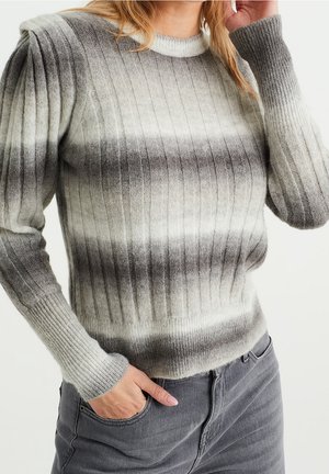 Vrouw draagt een geribde, grijs-wit verloop trui met lange mouwen, gecombineerd met grijze spijkerbroek, hand in zak, binnen.