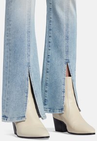 Jeans sfilati azzurri chiaro con aperture laterali, abbinati a stivali alla caviglia crema con punta appuntita e tacco a blocco nero.