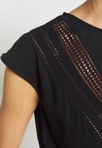 Gros plan d'un haut noir sans manches avec des panneaux en dentelle décorative sur l'épaule et le haut de la poitrine sur fond clair.