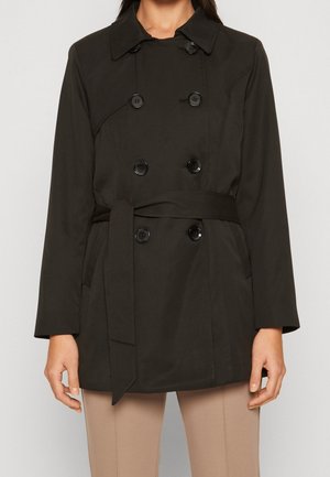 Kvinde iført en sort dobbeltradet trenchcoat med knapper og bælte i taljen, kombineret med beige bukser, stående mod en ensfarvet baggrund.