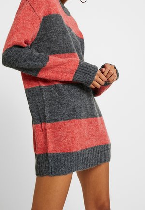 Le mannequin porte un long pull en maille duveteux à larges rayures corail et gris anthracite et se tient debout, les mains jointes, devant un fond uni.