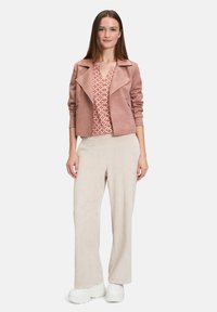 Vaaleanpunainen tekosuede-takki, jossa on leveä kaulus, kuvioitu paita ja beige kuristikankaiset väljät housut, yhdistettynä leveäpohjaisiin valkoisiin kenkiin.