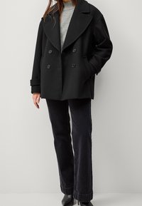 Abrigo negro de doble botonadura con grandes solapas, hecho de tela gruesa. Llevado sobre un suéter gris, combinado con jeans oscuros y botas de tacón.