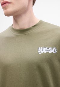 Suur lähedus pildil, kus on inimene, kellel on seljas oliivroheline särk, mille rinnal on valge stiliseeritud "HUGO" logo.