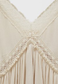 Cremfarbendes Kleidungsstück mit zarter Spitzenbordüre am Ausschnitt und einem gefalteten Design. Texturiert mit einem sanften, fließenden Aussehen.