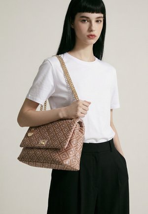 Jonge vrouw met zwart haar, die een wit t-shirt en zwarte broek draagt, houdt een bruine handtas met patroon en een gouden kettingriem over haar schouder.