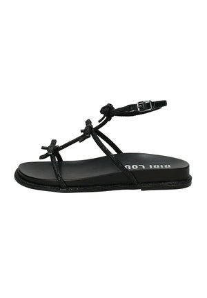 Sandalia plana negra con tiras finas, detalles anudados, hebilla en el tobillo y suela ligeramente elevada, mostrando texto parcial de la marca en blanco en la plantilla.