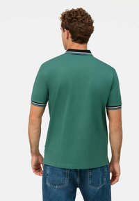 Polo verde con una trama uniforme, caratterizzato da un colletto blu navy e accenti a strisce sulle maniche. Indossato con jeans in denim blu.