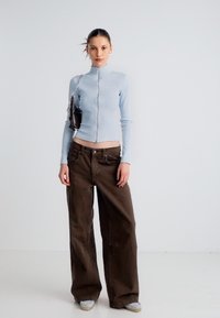 Ελαφρύ μπλε ριμπ ζακέτα με φερμουάρ σε συνδυασμό με φαρδιά καφέ παντελόνια. Το μοντέλο κρατά μια μικρή μαύρη τσάντα και φορεί ανοιχτό γκρι αθλητικά παπούτσια.