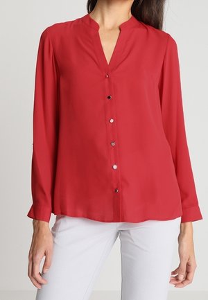 Femme portant un chemisier rouge à manches longues boutonné avec un col en V et un pantalon blanc, debout sur un fond neutre.