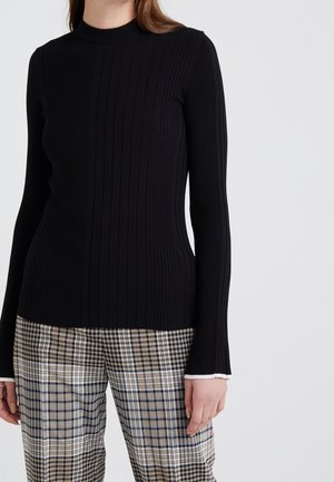 Kobieta ubrana w przylegającą czarną prążkowaną bluzkę z długim rękawem oraz beżowe, czarno-biało-beżowe spodnie w kratę, stojąca na tle jednolitego jasnego tła.