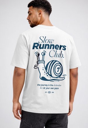 Man wearing white t-shirt with "Slow Runners Club" text and a cartoon snail on the back, paired with dark jeans.