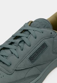 Detailní záběr na šedou tenisku Reebok s texturovanými tkaničkami, viditelným logem značky na boku a materiálem podobným semiši.