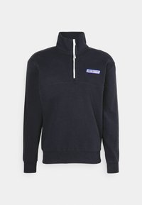 Sudadera de color navy con cuello alto y cierre de media cremallera en la parte delantera. Presenta una cremallera blanca y un parche de logo en el pecho. Textura de tela suave.