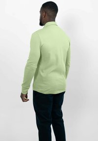 Langsleeve, lichtgroen poloshirt met een kraag en een aangesloten ontwerp, gedragen met donkerblauwe broeken, gezien vanaf de achterkant tegen een witte achtergrond.
