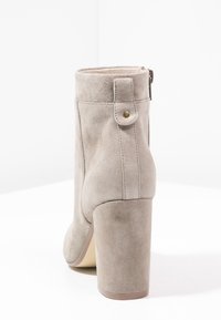 Stivale alla caviglia in suede beige con tacco a blocco alto, zip posteriore e punta arrotondata. Presenta un pannello laterale cucito e un bottone decorativo.