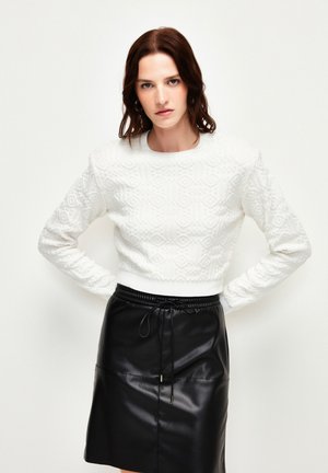 Pull en maille texturée blanche avec des motifs géométriques, associé à une jupe en cuir noir avec une taille à cordon.
