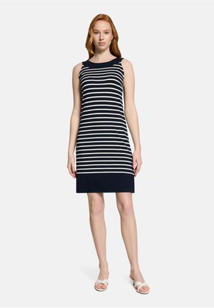 Femme portant une robe sans manches à rayures horizontales bleu marine et blanches jusqu'aux genoux avec un ourlet bleu marine et des sandales blanches, debout sur un fond uni.