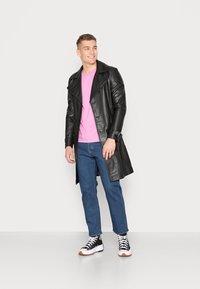 Casaco trench de couro preto com grandes lapelas, combinado com uma t-shirt rosa, jeans azuis e sapatilhas pretas com atacadores brancos e solas laranja.