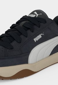 Μαύρο-άσπρο αθλητικό παπούτσι Puma με μαύρα κορδόνια, λευκό λογότυπο στο πλάι και στη γλώσσα, μπεζ ενδιάμεση σόλα και καφέ εξωτερική σόλα.