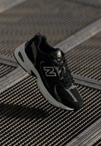 Μαύρο και γκρι αθλητικό παπούτσι New Balance με λευκή σόλα τοποθετημένο σε μεταλλική επιφάνεια πλέγματος, σε γωνία για να δείξει πλευρικές και επάνω όψεις.