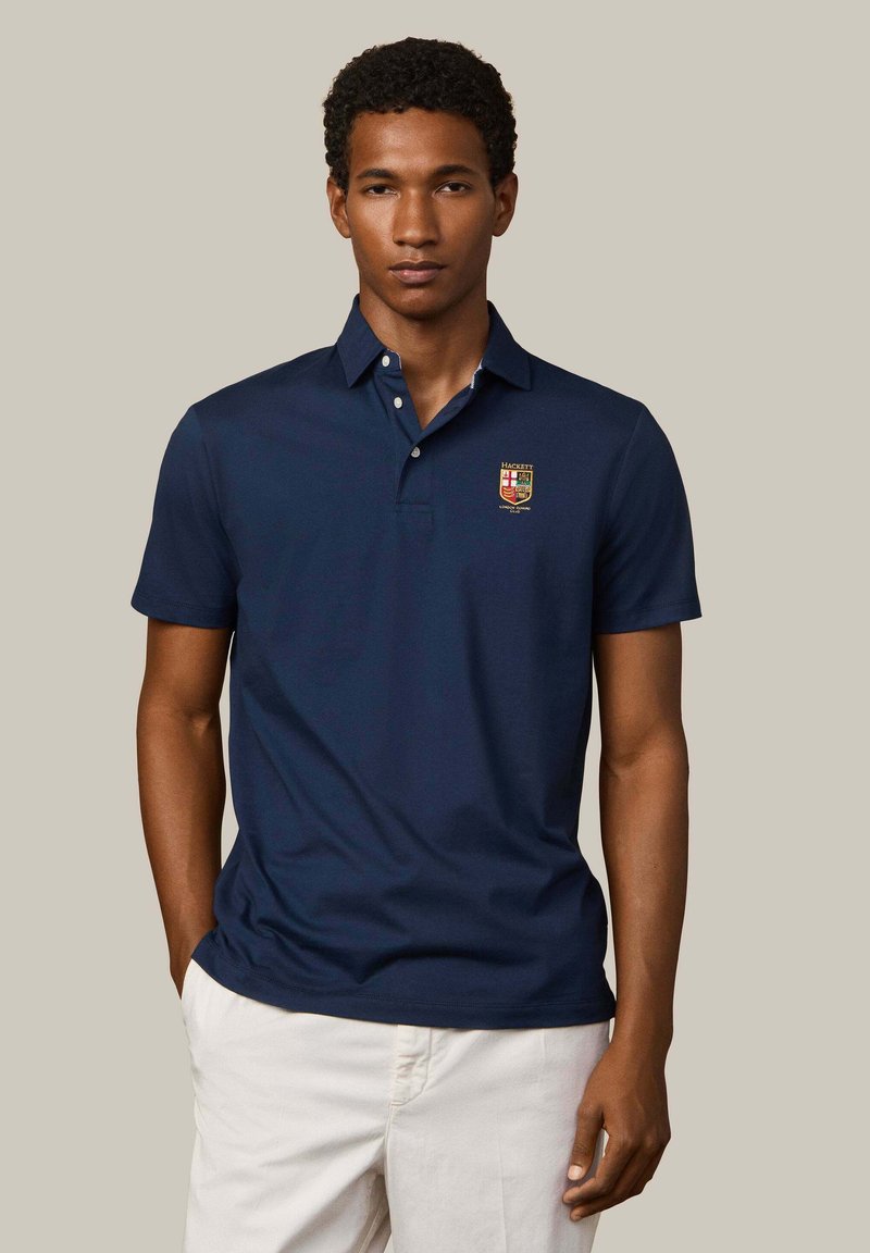 Hombre con camisa polo azul marino con emblema bordado y pantalones blancos, de pie con una mano en el bolsillo sobre un fondo liso.