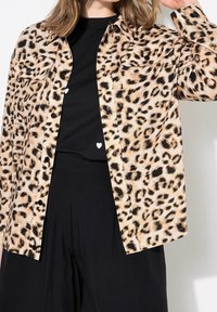 Leopard-print shirt in tan and black fabric, featuring button closures, two chest pockets, and a relaxed fit, layered over a black top.