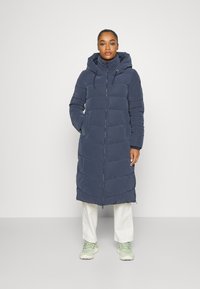 Mørkeblå lang pufferjakke med hætte, der har vandret quiltede sektioner, lynlåsåbning og sidelommer. Bæret over lyse bukser og sneakers.