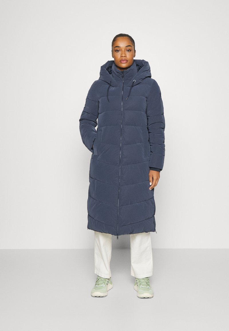 Mørkeblå lang pufferjakke med hætte, der har vandret quiltede sektioner, lynlåsåbning og sidelommer. Bæret over lyse bukser og sneakers.